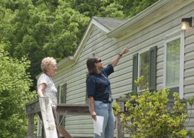 Two women make your roof ready for summers by standing on a ladder in front of a house.