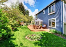 A backyard with a blue house and green grass, requiring siding replacement.